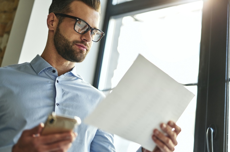 Een data-analist met een bril is aan het multitasken, houdt een stuk papier vast en kijkt op zijn telefoon naar informatie.